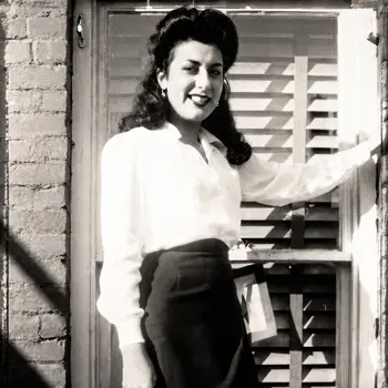 Black-and-white photograph of a smiling young woman in a white blouse standing by a window