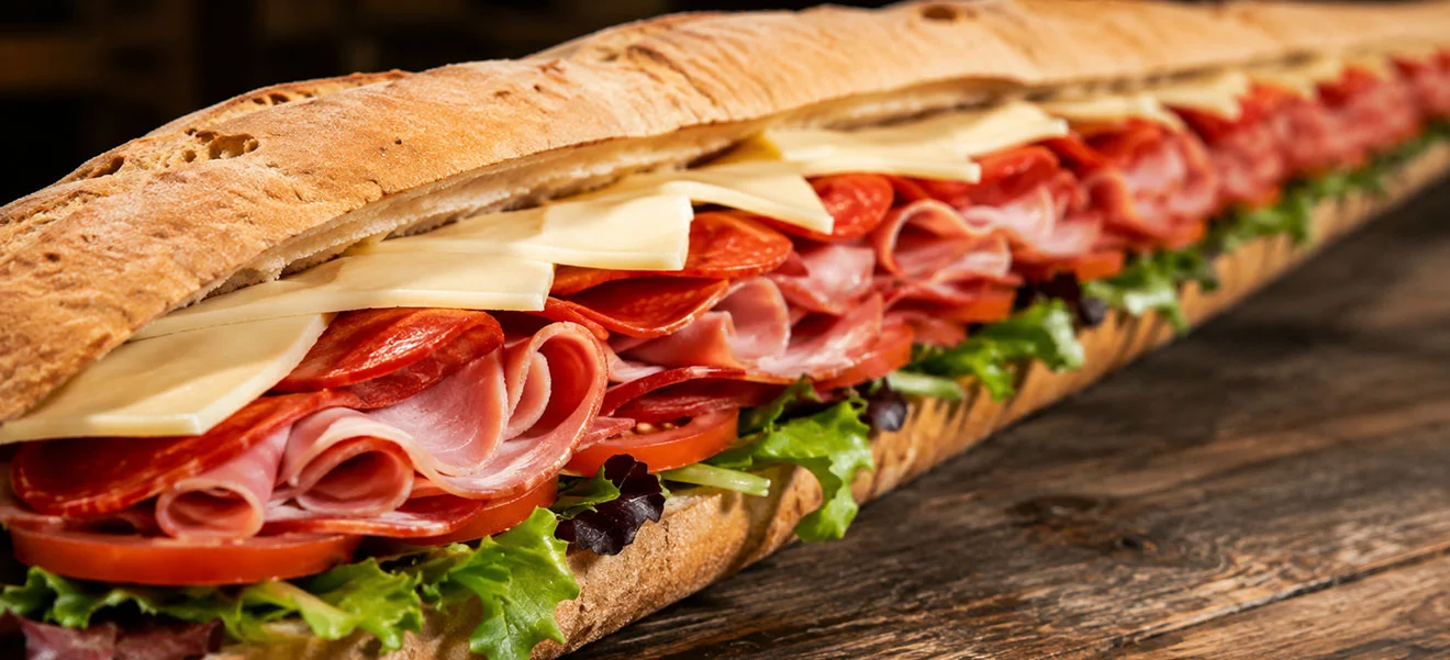 A long Italian sub layered with ham, pepperoni, provolone, tomato and lettuce on a wooden table